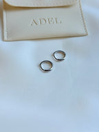 Silver hoop earrings on a white surface with a beige pouch labeled 'ADEL' in the background.
