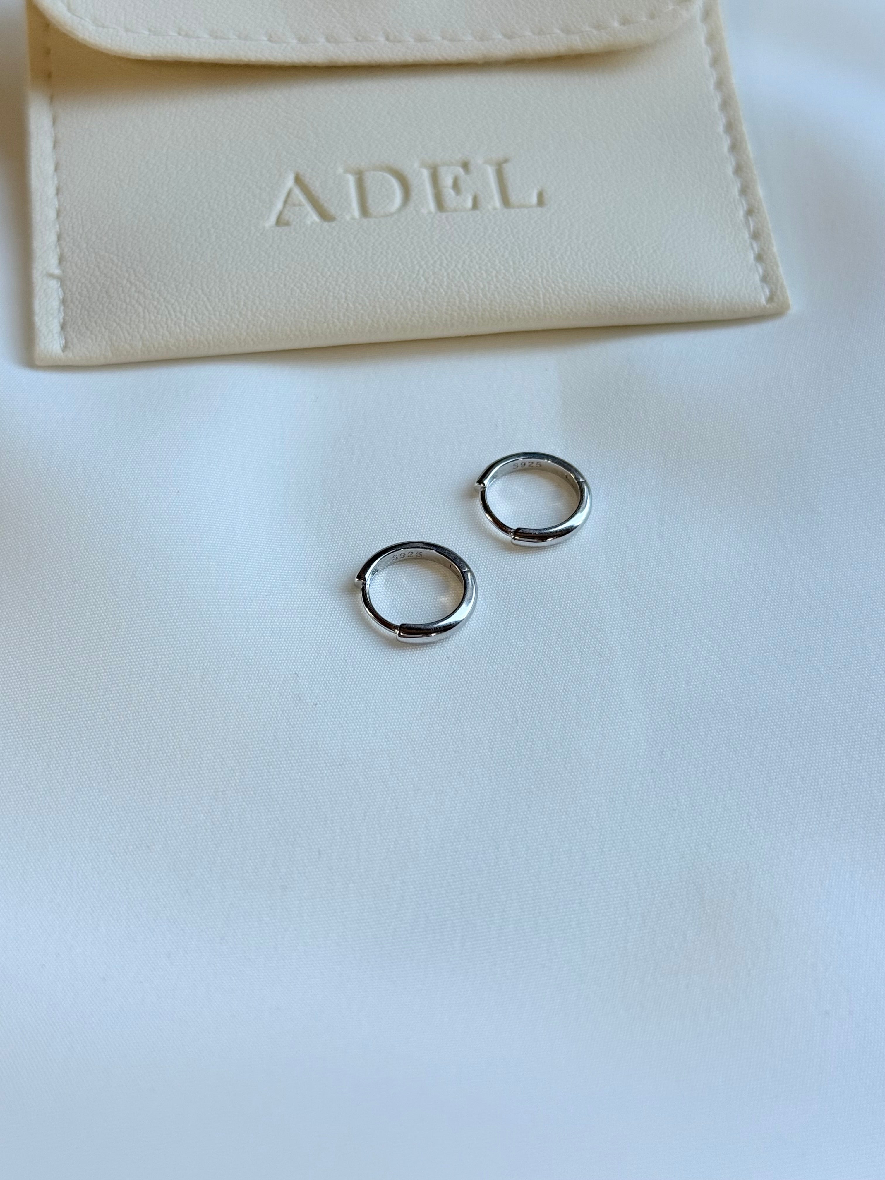 Silver hoop earrings on a white surface with a beige pouch labeled 'ADEL' in the background.
