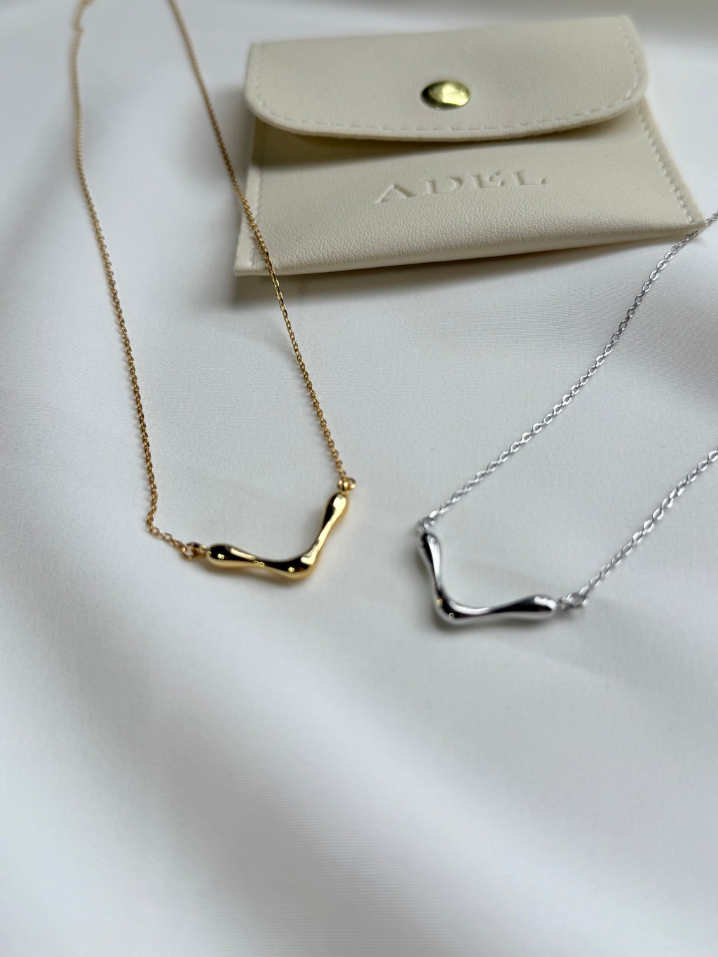 Gold and silver necklaces on a white surface with a beige pouch labeled 'ADEL'.