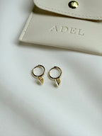Gold hoop earrings with leaf-shaped ends on a white surface next to a beige pouch with 'ADEL' branding.