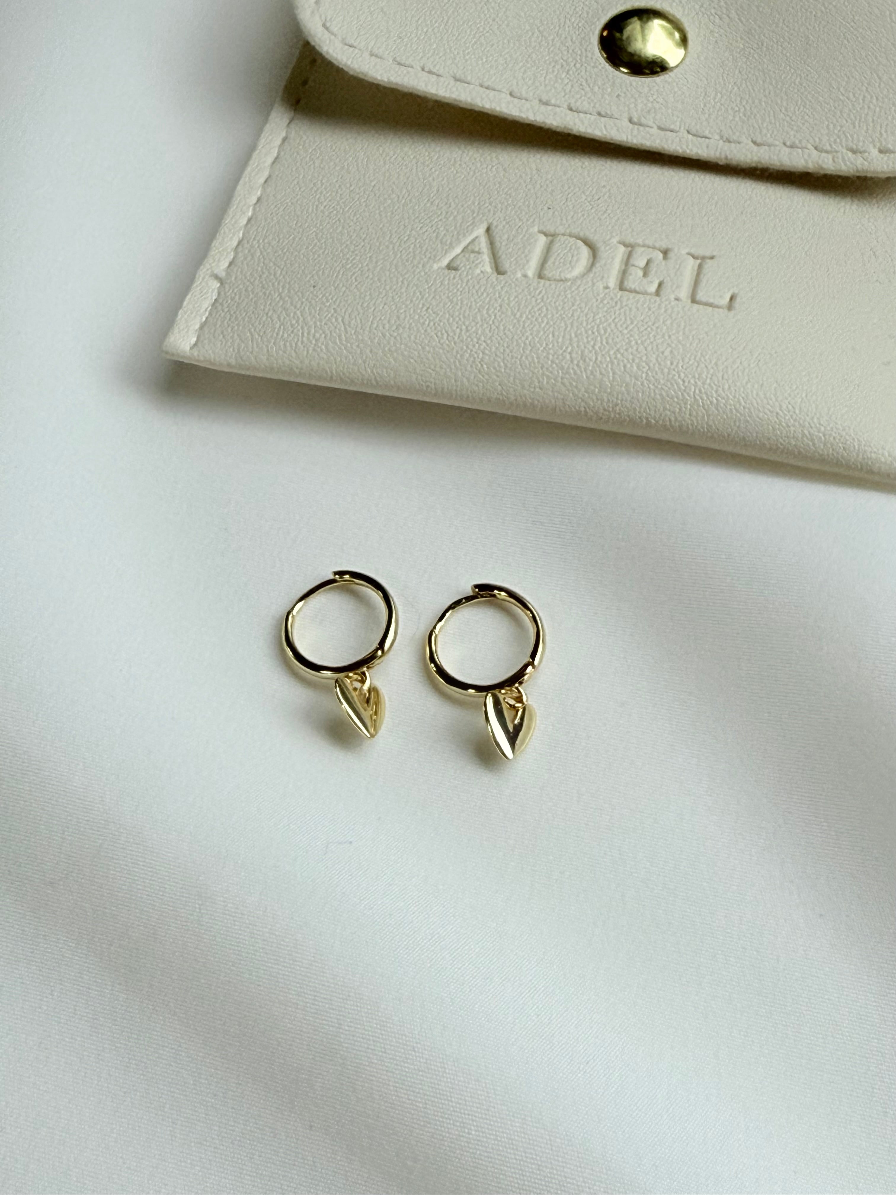 Gold hoop earrings with leaf-shaped ends on a white surface next to a beige pouch with 'ADEL' branding.