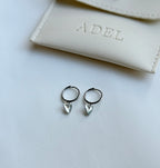 Silver hoop earrings with heart-shaped charms on a white surface next to a beige pouch with 'A.DEL' branding.