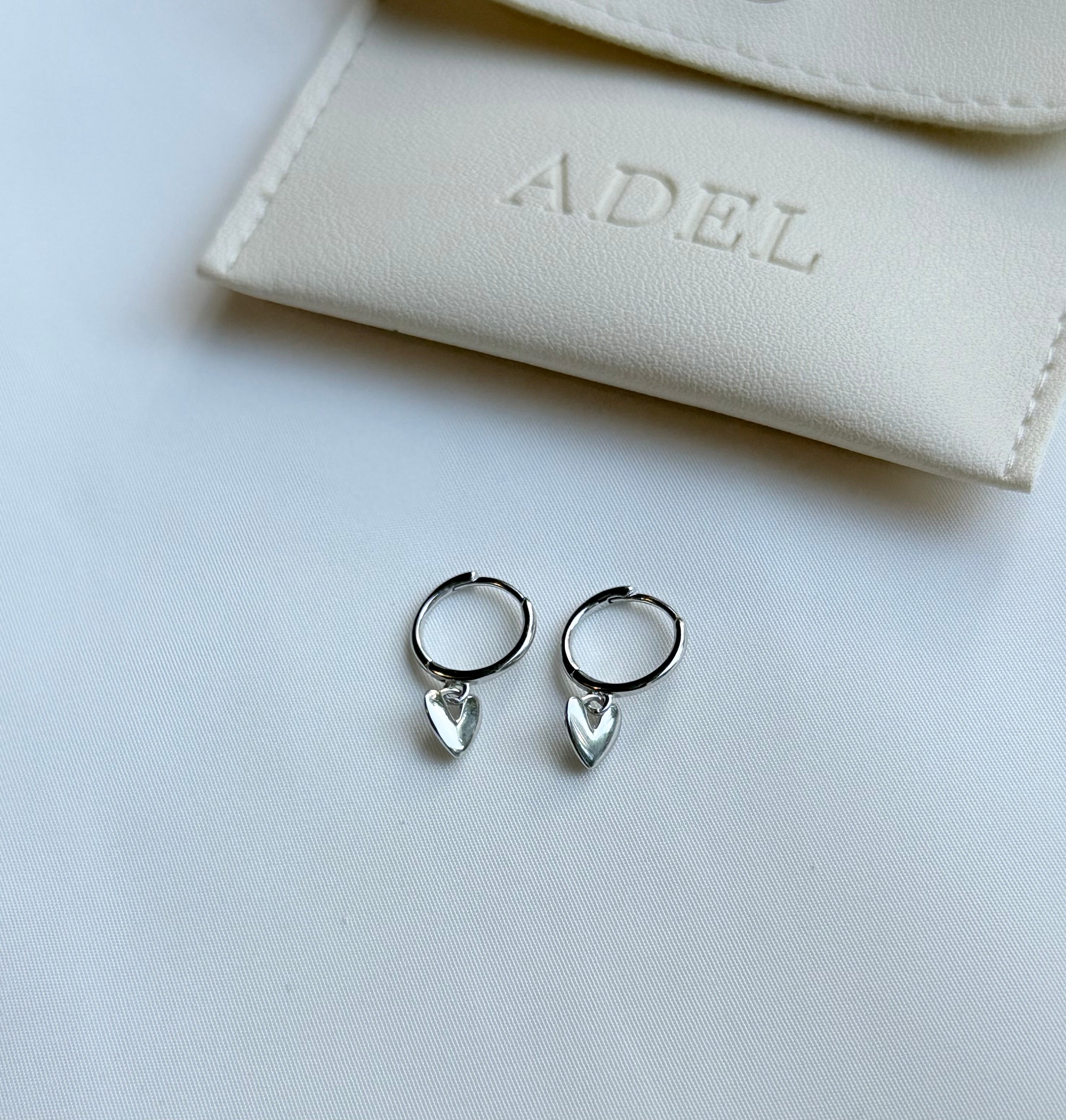 Silver hoop earrings with heart-shaped charms on a white surface next to a beige pouch with 'A.DEL' branding.