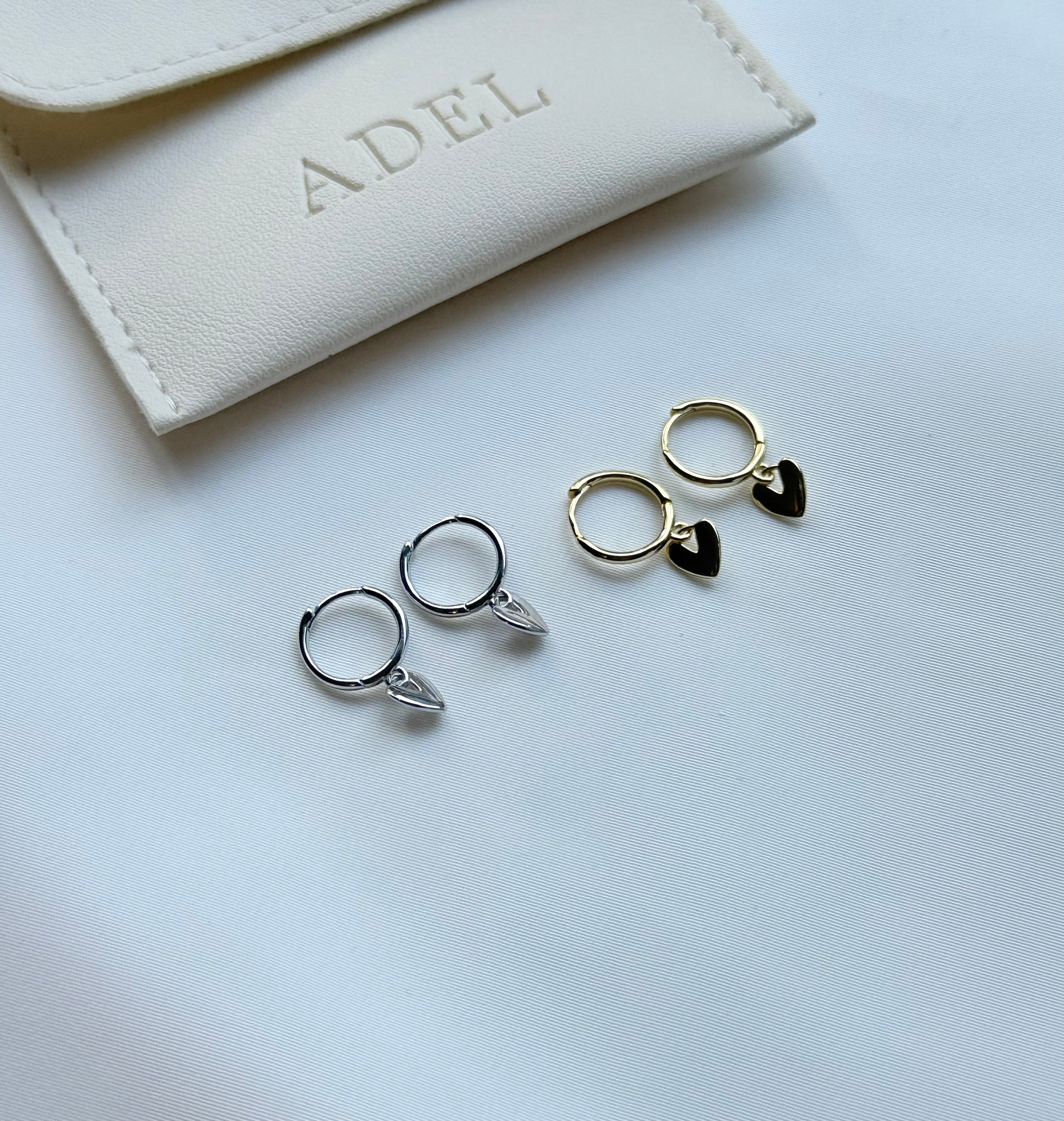 Two pairs of hoop earrings with leaf details on a light gray surface, with a beige jewelry pouch labeled 'ADEL' in the background.