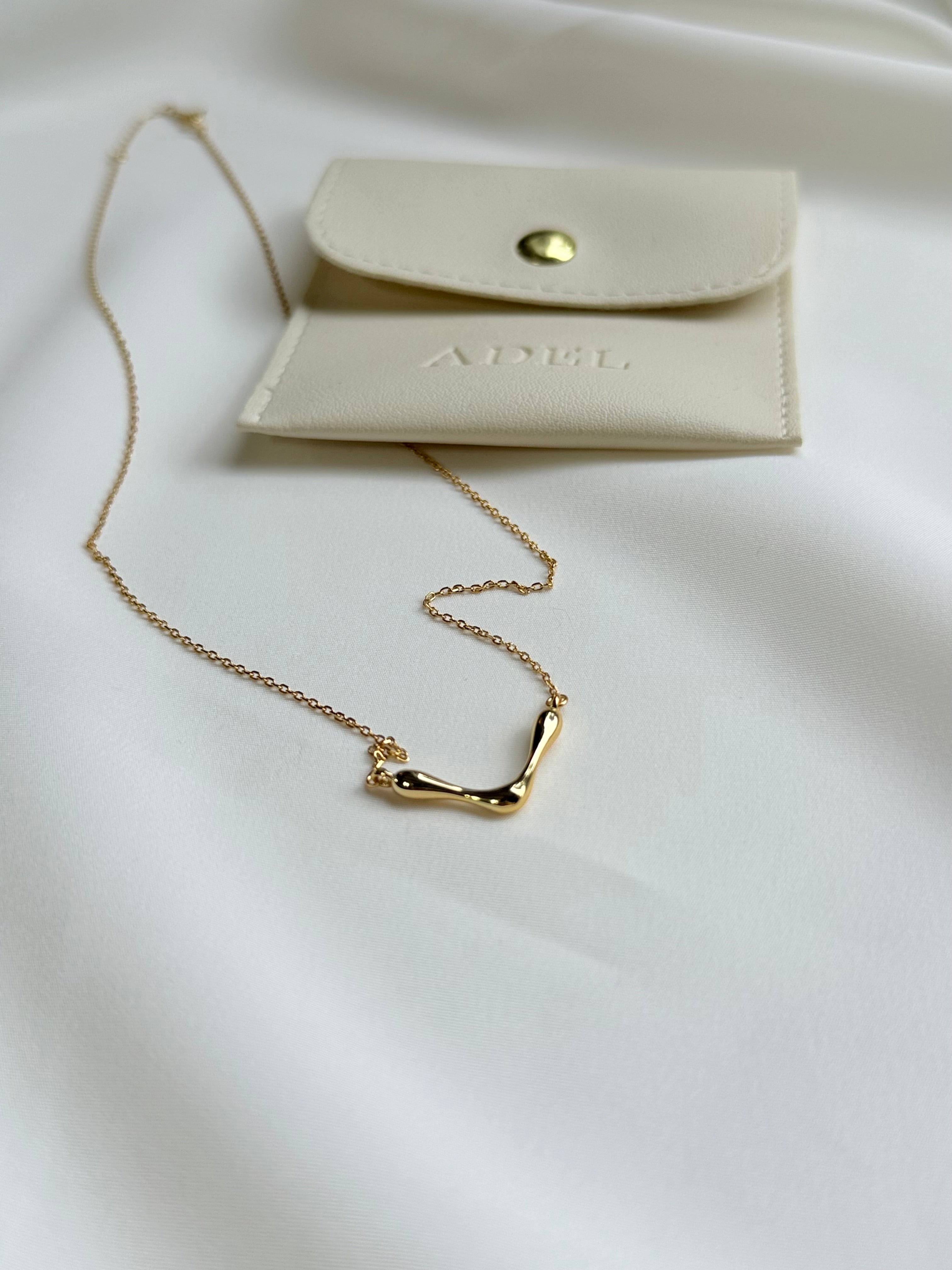 Gold necklace on a white surface with a beige card featuring 'ADEL' branding.