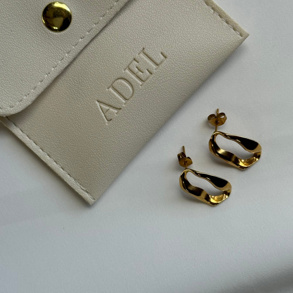 Gold hoop earrings next to a beige jewelry pouch with 'ADEL' branding on a light gray background
