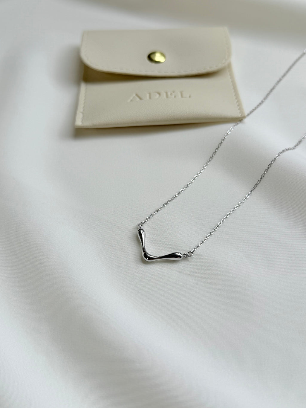 Silver necklace on a white surface with a beige pouch labeled 'ADEL'.