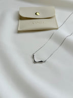 Silver necklace on a white surface with a beige pouch labeled 'ADEL'.