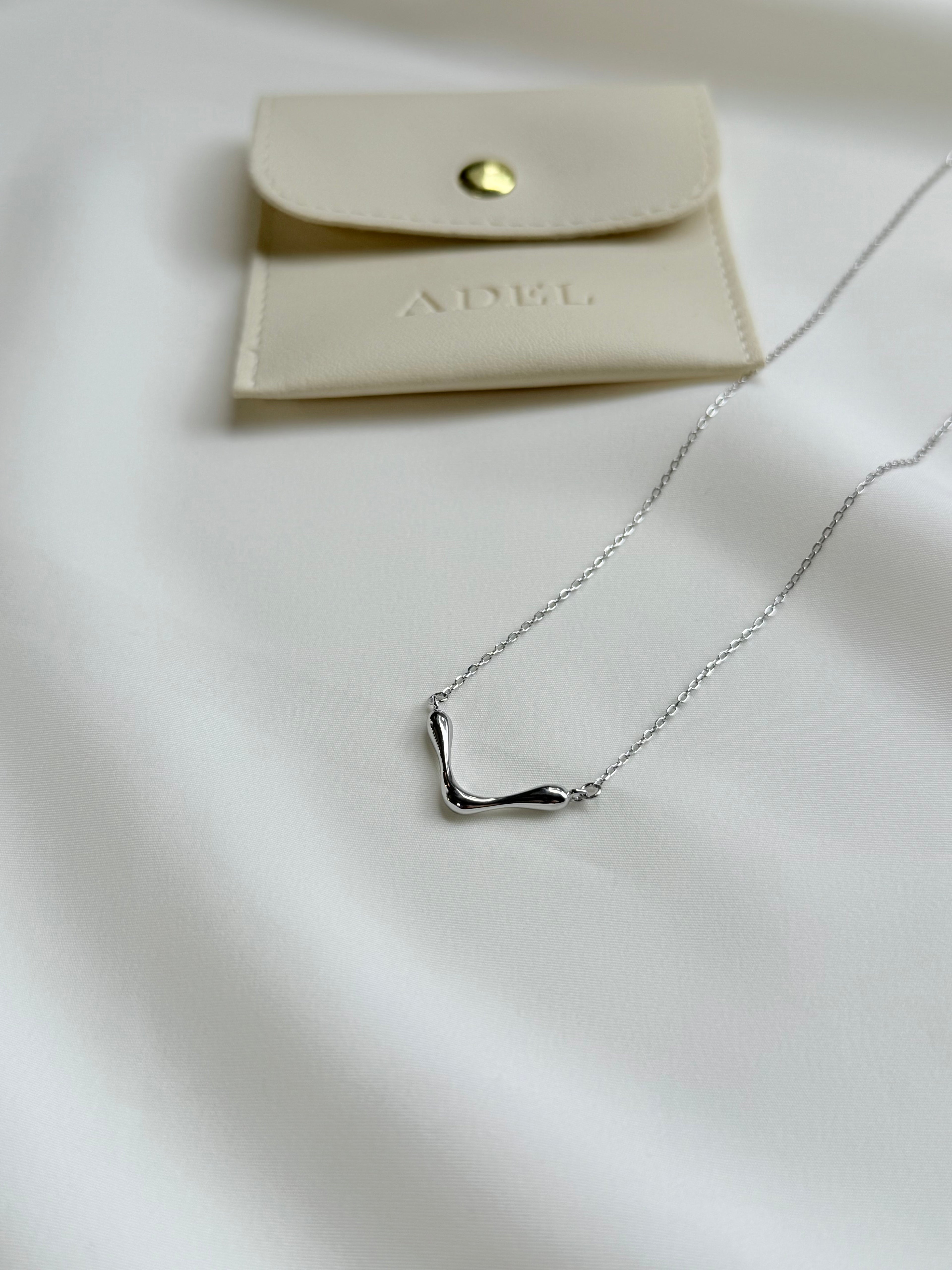 Silver necklace on a white surface with a beige pouch labeled 'ADEL'.