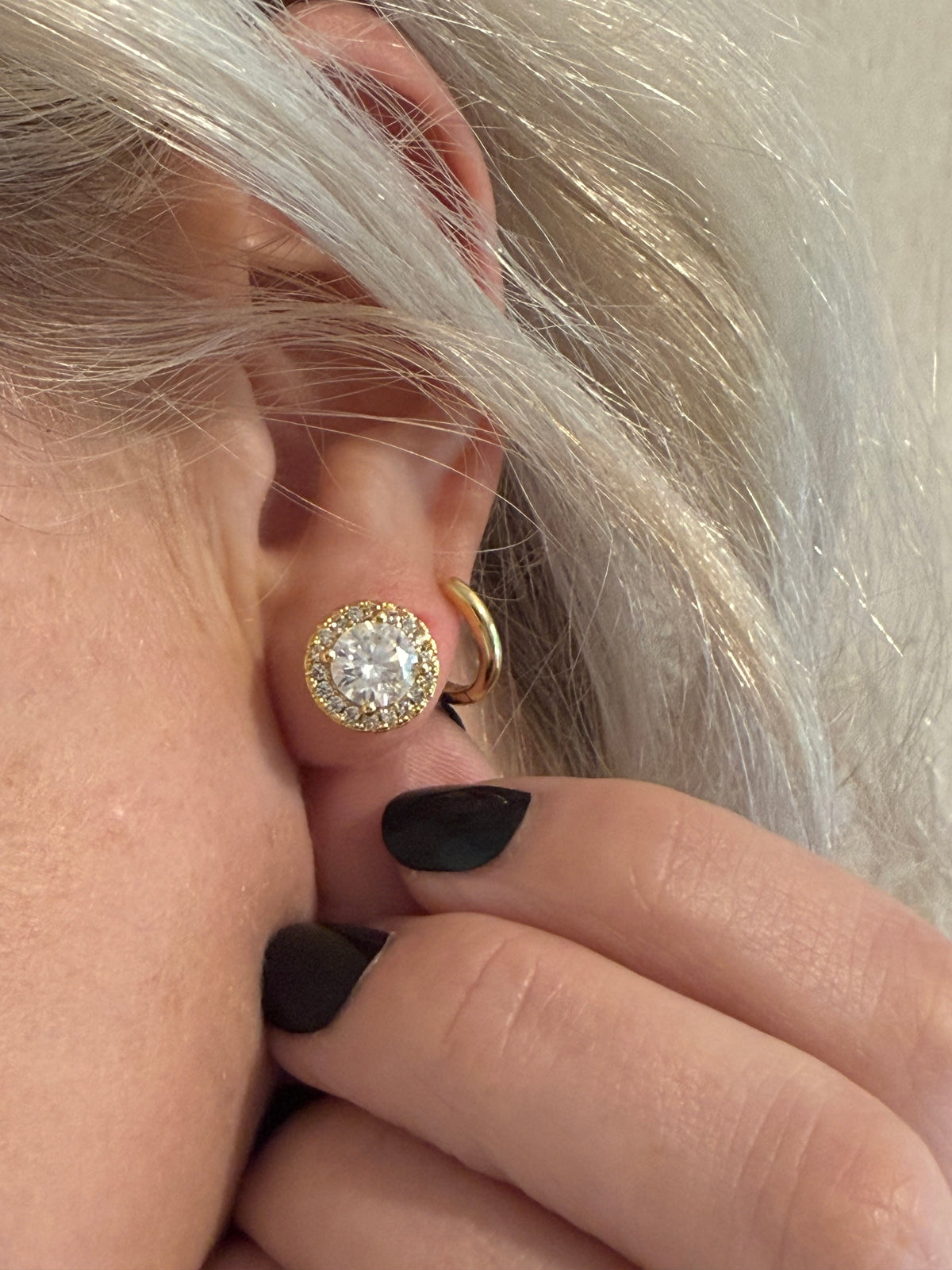 A close-up of a person's ear wearing a gold hoop earring with a round clear gemstone surrounded by a halo of smaller clear stones, with fingers touching the ear.