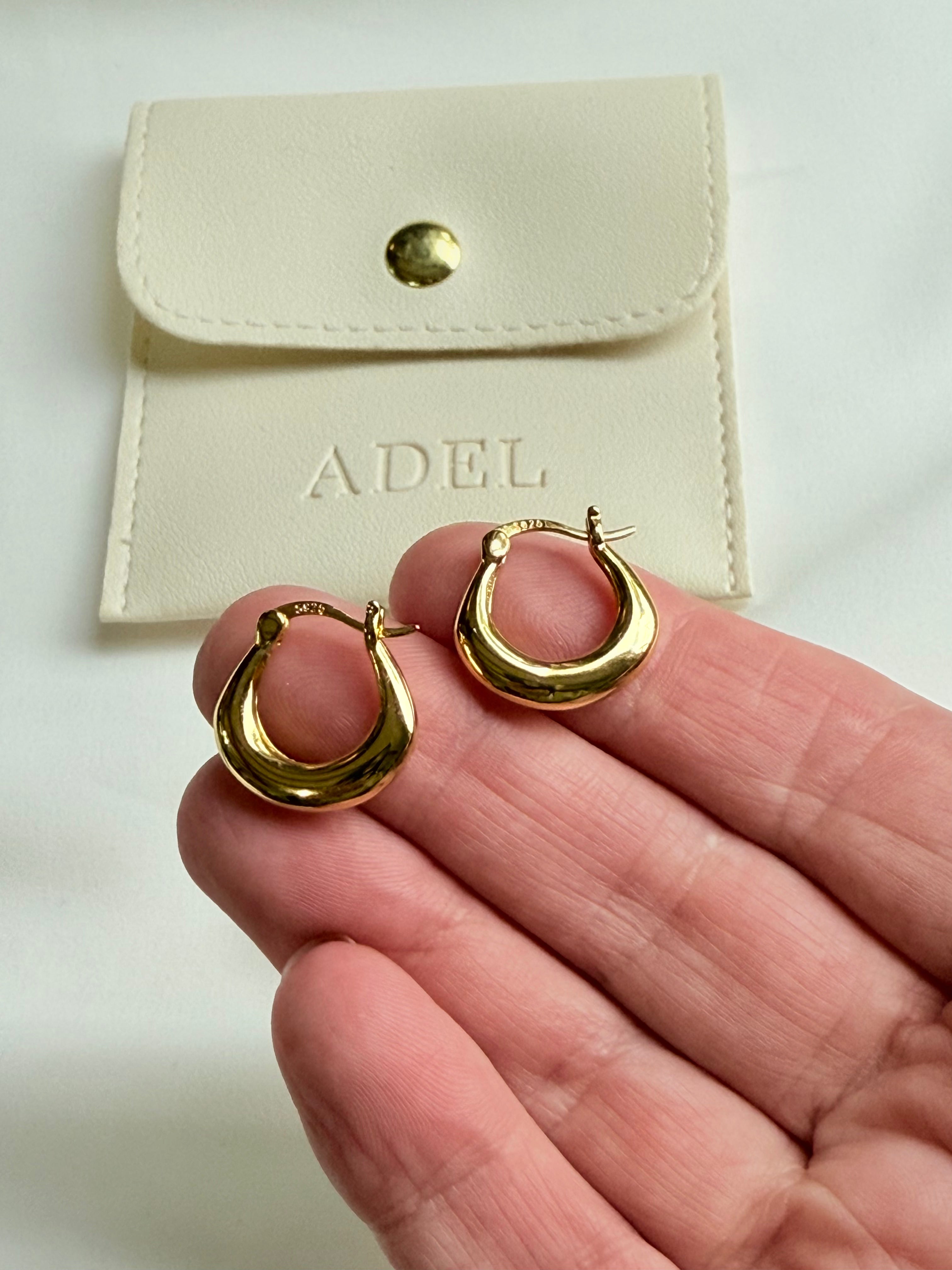 Gold hoop earrings held in a hand with a beige pouch labeled 'ADEL' in the background.