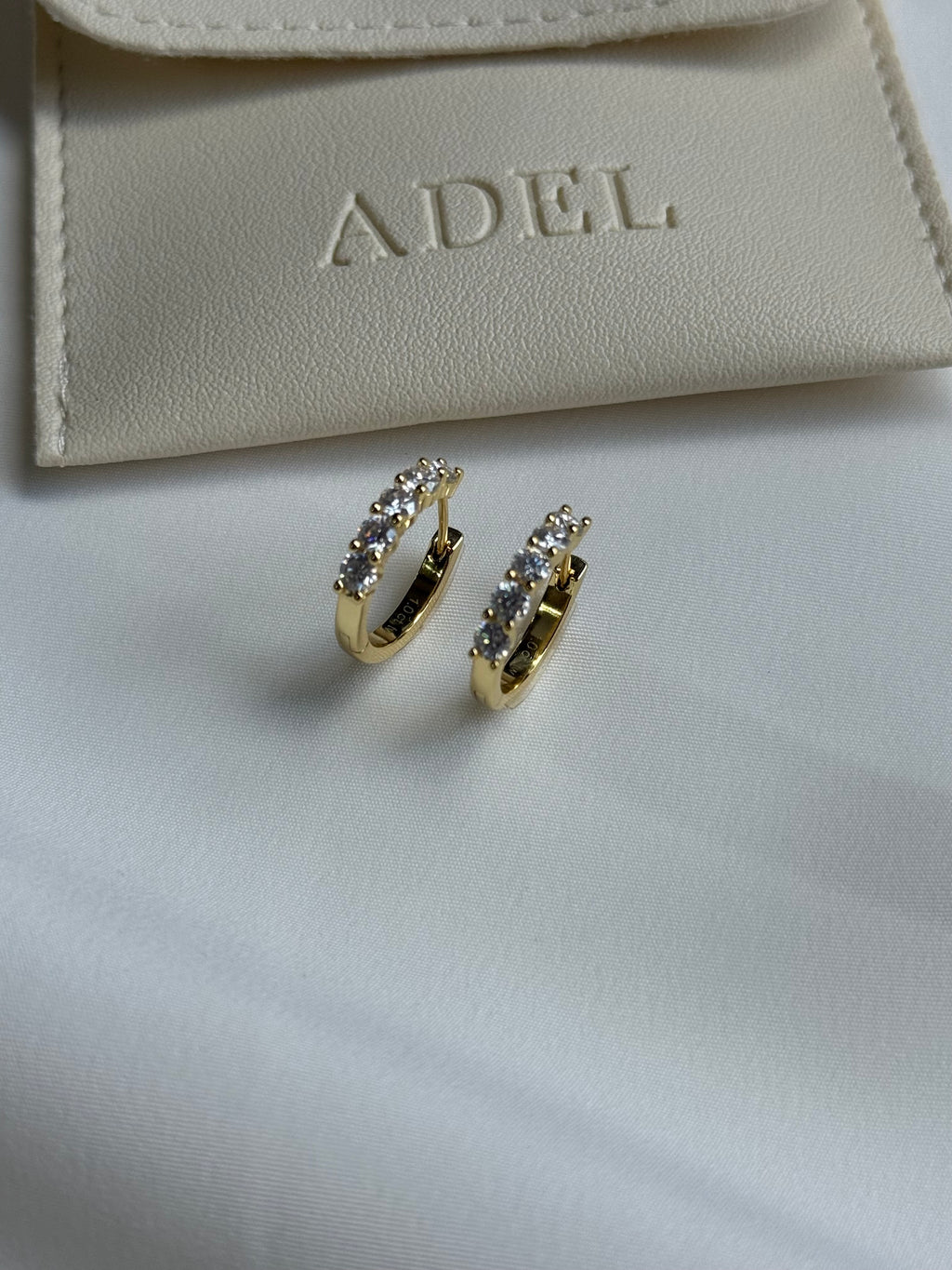 Gold hoop earrings with embedded stones on a beige surface with 'ADEL' branding.