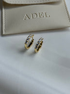 Gold hoop earrings with embedded stones on a beige surface with 'ADEL' branding.