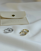 Two pairs of earrings, one silver and one gold, on a white surface with a beige pouch in the background.