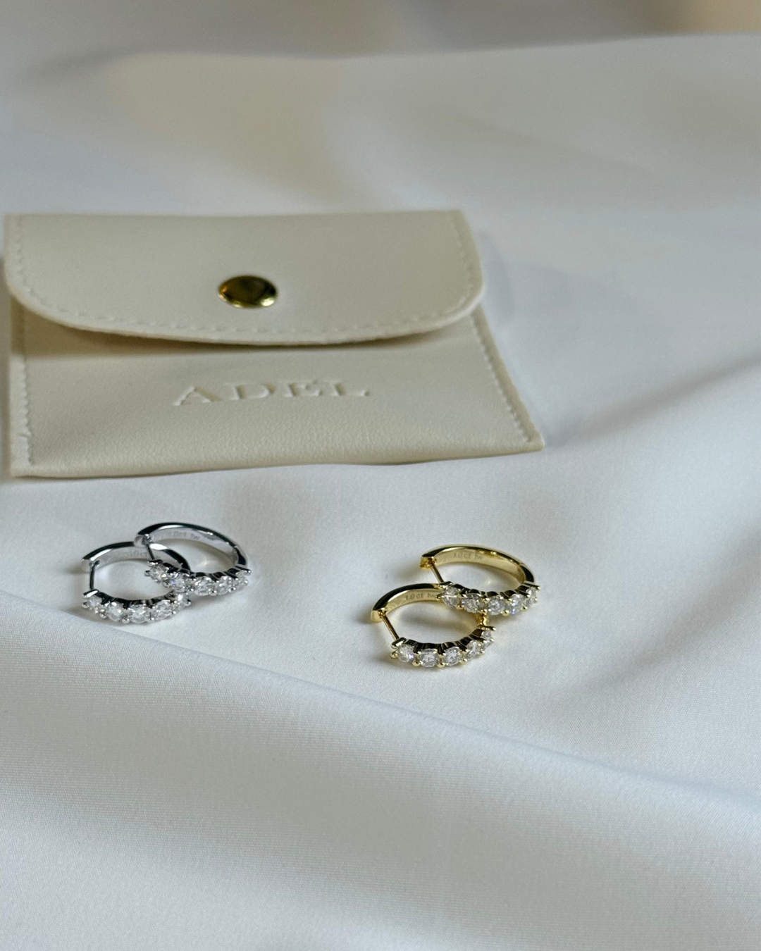 Two pairs of earrings, one silver and one gold, on a white surface with a beige pouch in the background.
