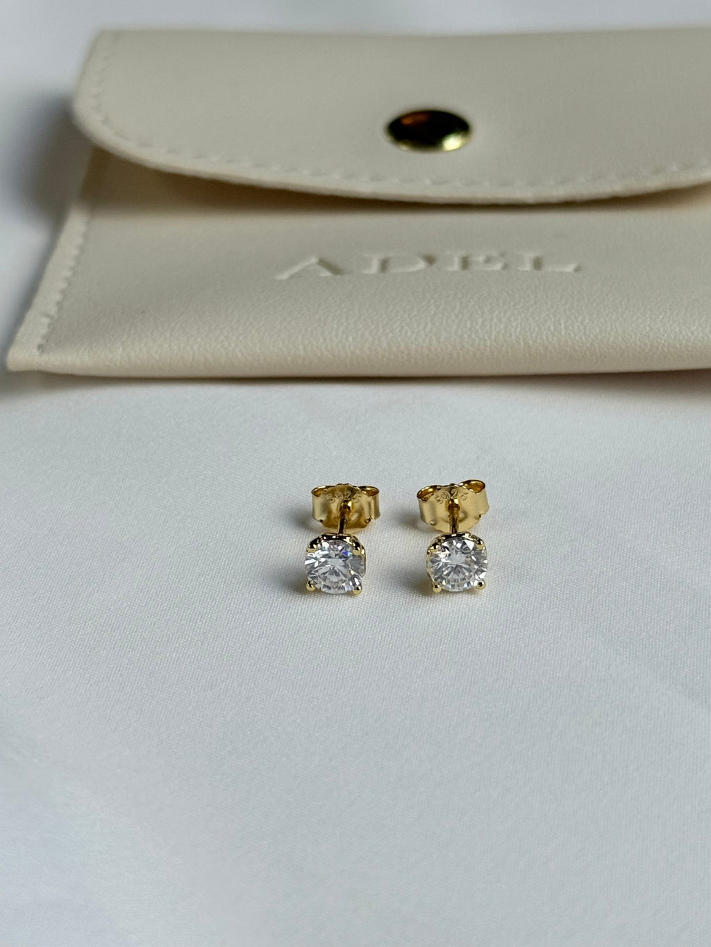 Gold and diamond stud earrings on a light gray surface with a beige pouch in the background.