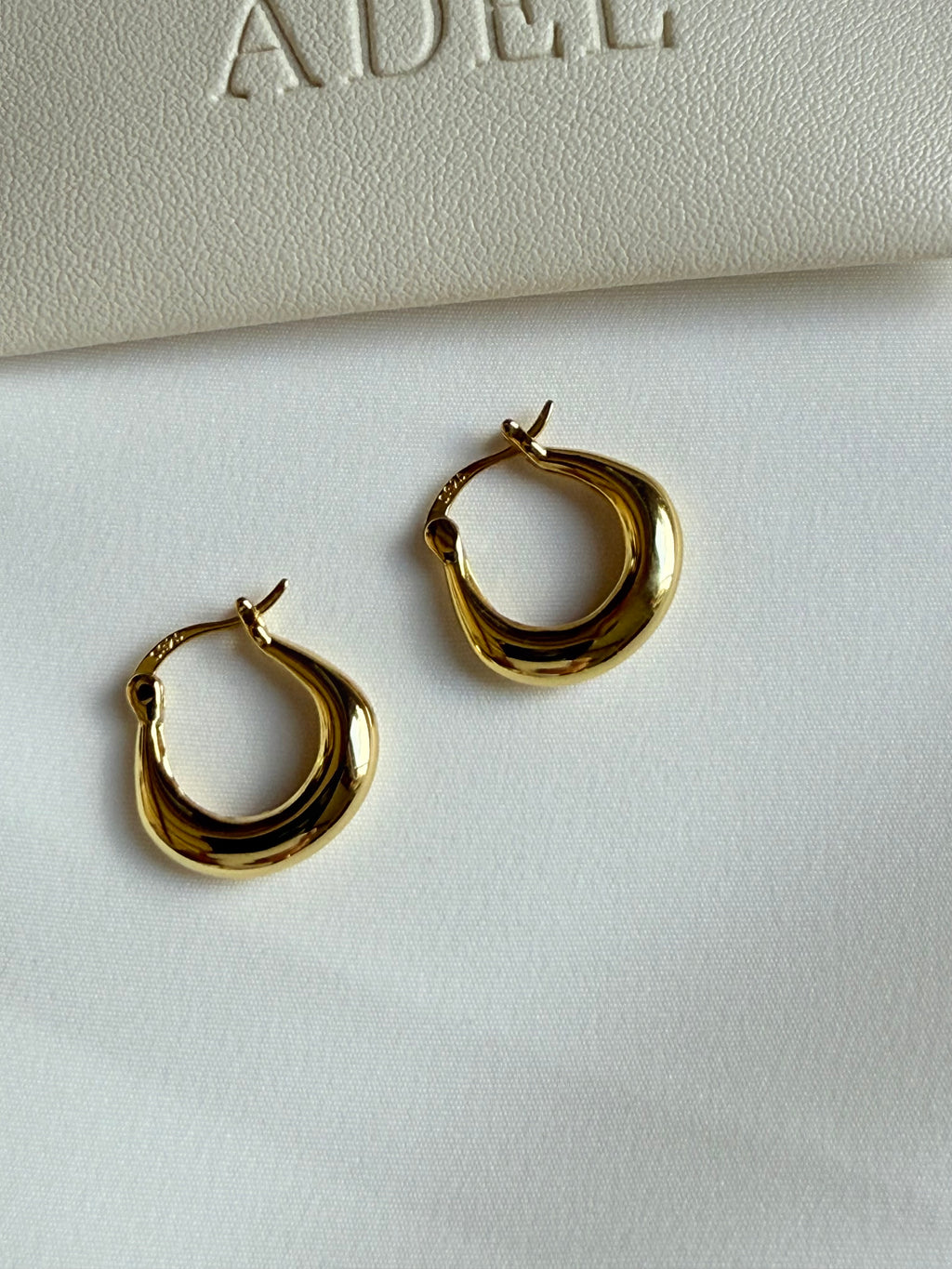 Gold hoop earrings on a white surface with 'ADEL' branding in the background.
