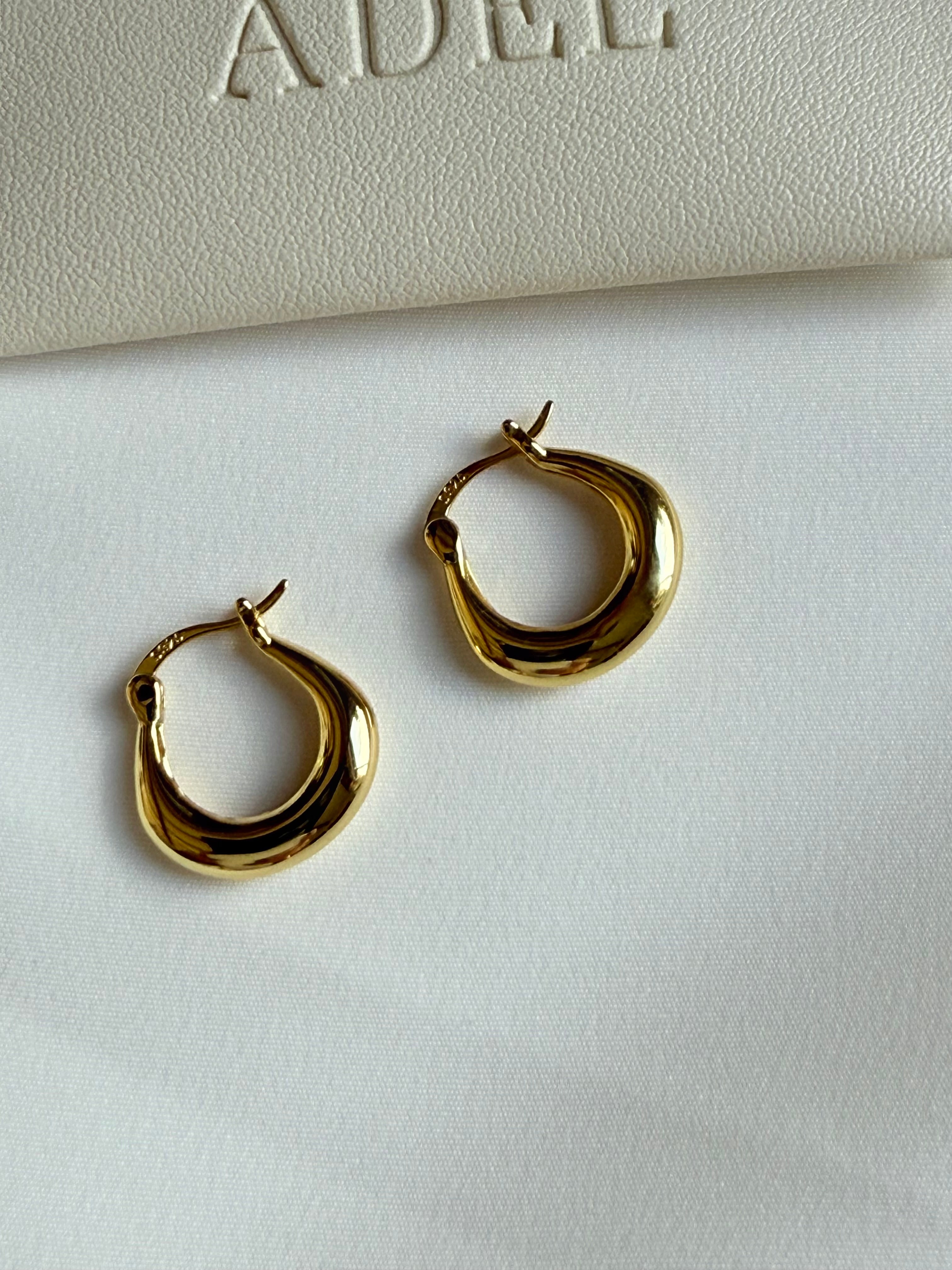Gold hoop earrings on a white surface with 'ADEL' branding in the background.