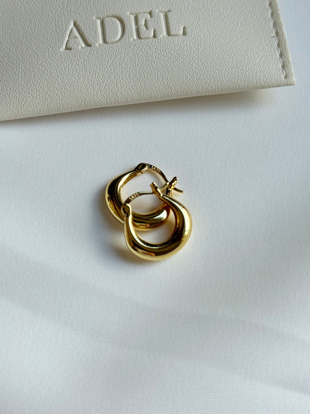 Gold hoop earrings on a white surface with 'ADEL' branding in the background.