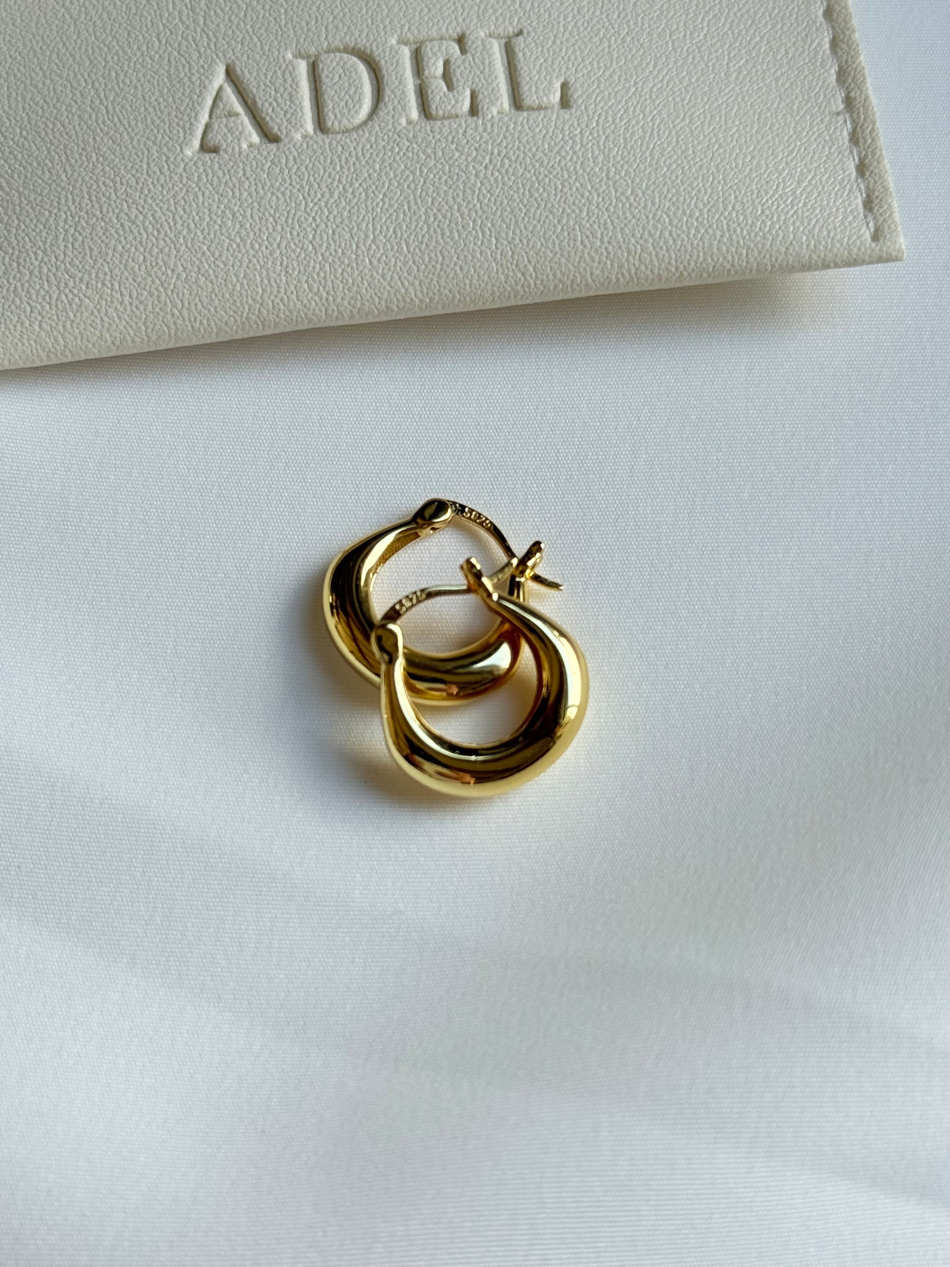 Gold hoop earrings on a white surface with 'ADEL' branding in the background.