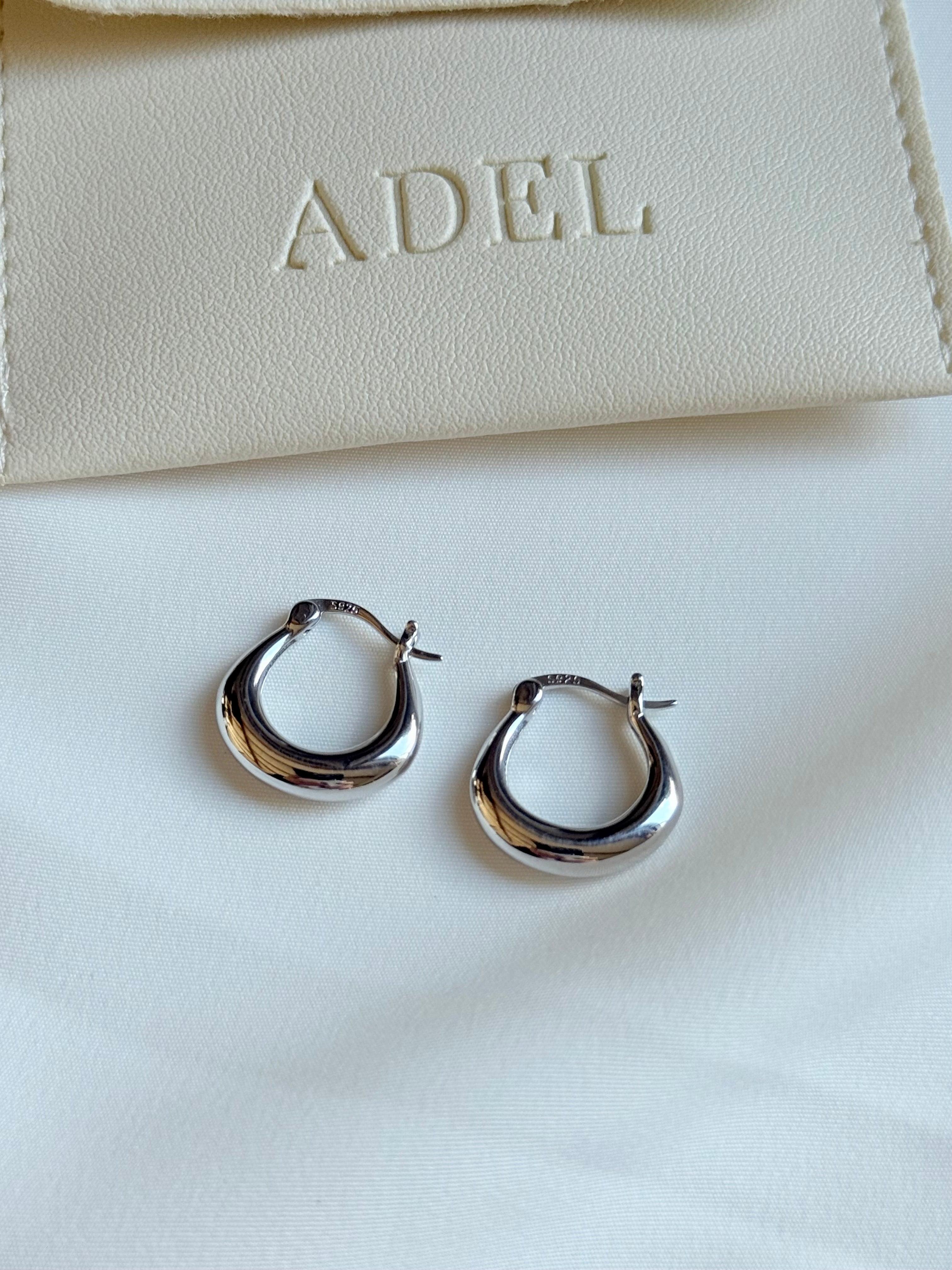 Silver hoop earrings on a white surface with a beige pouch labeled 'ADEL' in the background.