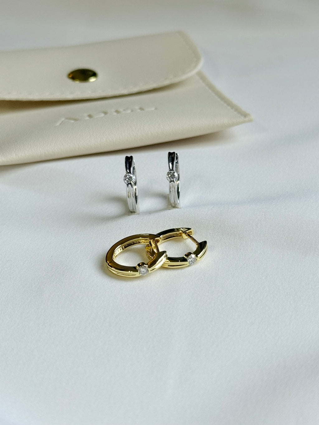 Gold and silver hoop earrings on a white surface with a beige pouch in the background.