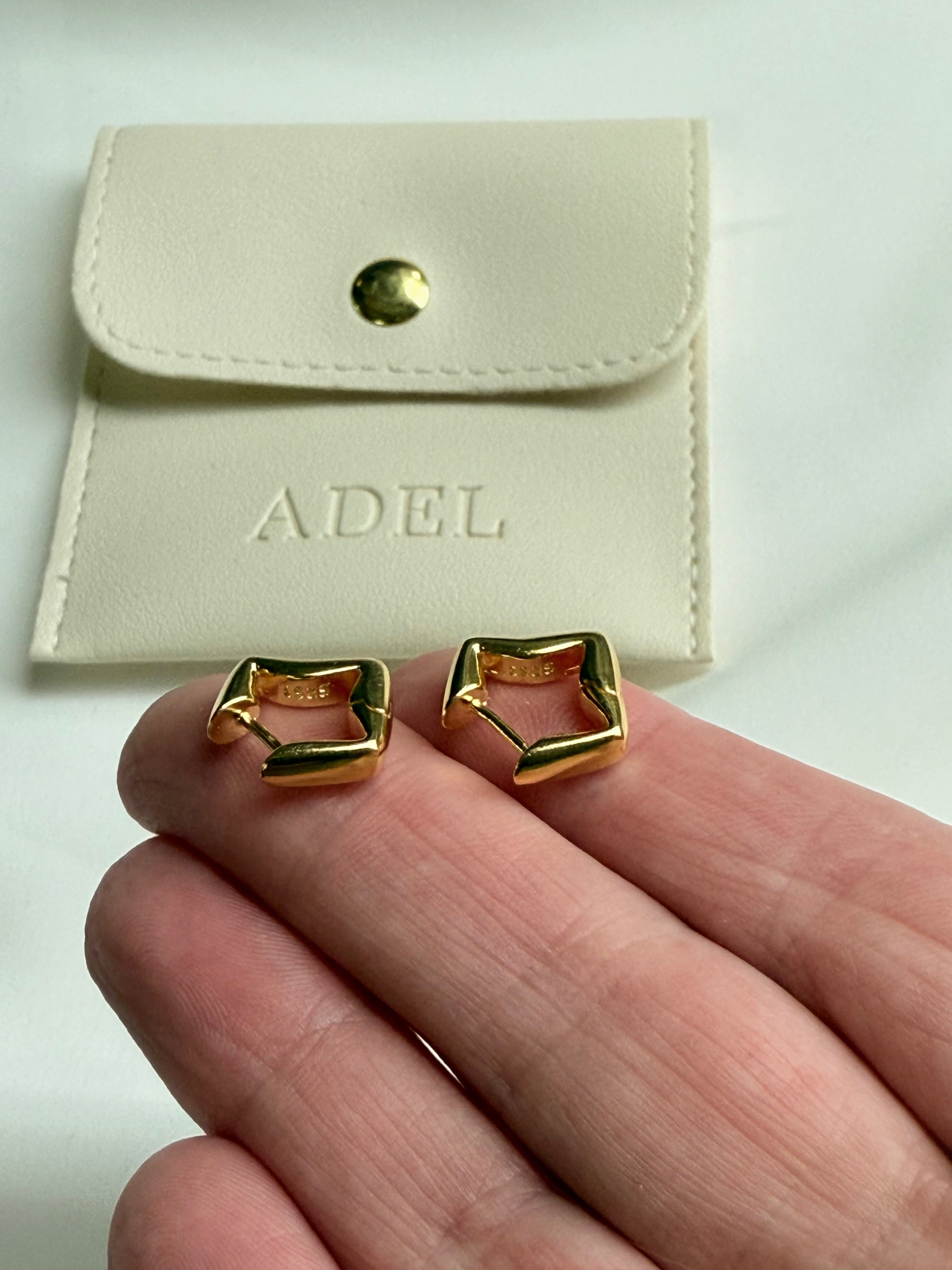 Gold earrings held between fingers with a beige pouch labeled 'ADEL' in the background.