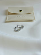 Pair of silver hoop earrings on a white surface with a beige jewelry pouch in the background.