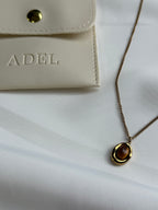 Necklace with a pendant on a white surface next to a beige pouch with 'ADEL' branding.